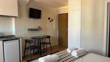 A compact hotel room with a kitchenette, a single bed with two rolled towels, a small dining table with two chairs, a TV mounted on the wall, and a wardrobe.
