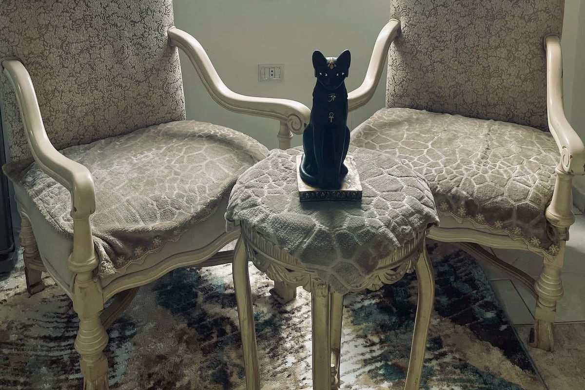 Dos sillones con una estatua decorativa de un gato entre ellos, un tapete estampado y vistas a las pirámides a través de una ventana.