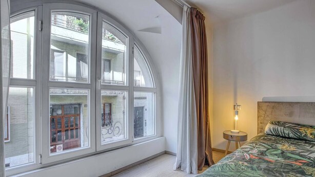 A bedroom with a large arched window, a bed with a floral bedspread, a bedside table with a lamp, and a view of the street outside. - DUOMO 10min - Lovely flat in the center (Milano)