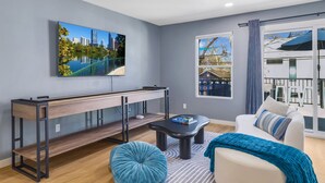 Un salon moderne avec un tableau représentant un paysage urbain, un banc en bois, un pouf bleu, un canapé blanc et une vue sur un balcon.