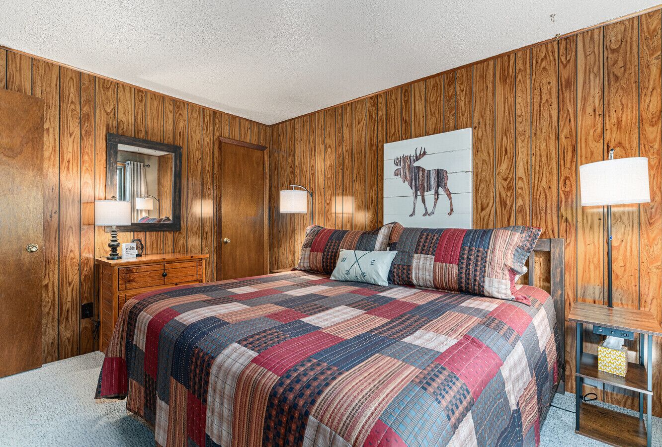 A bedroom with wooden paneling, a large bed with a colorful quilt, two bedside lamps, a wooden dresser, and a framed picture on the wall.