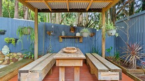 An outdoor wooden pergola with a built-in bench and table, surrounded by a blue fence and various potted plants.