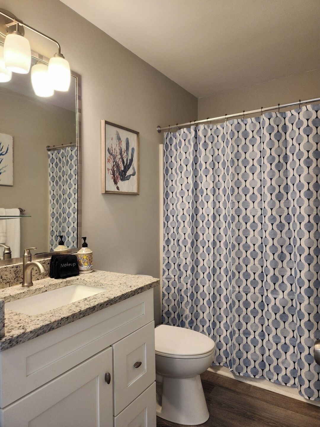 Une salle de bain avec un meuble-lavabo, un évier et un miroir. On y trouve un rideau de douche à motifs géométriques, des toilettes et une œuvre d’art encadrée accrochée au mur.
