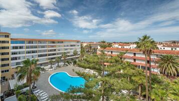A hotel with a swimming pool surrounded by palm trees and lounge chairs, overlooking a coastal area.