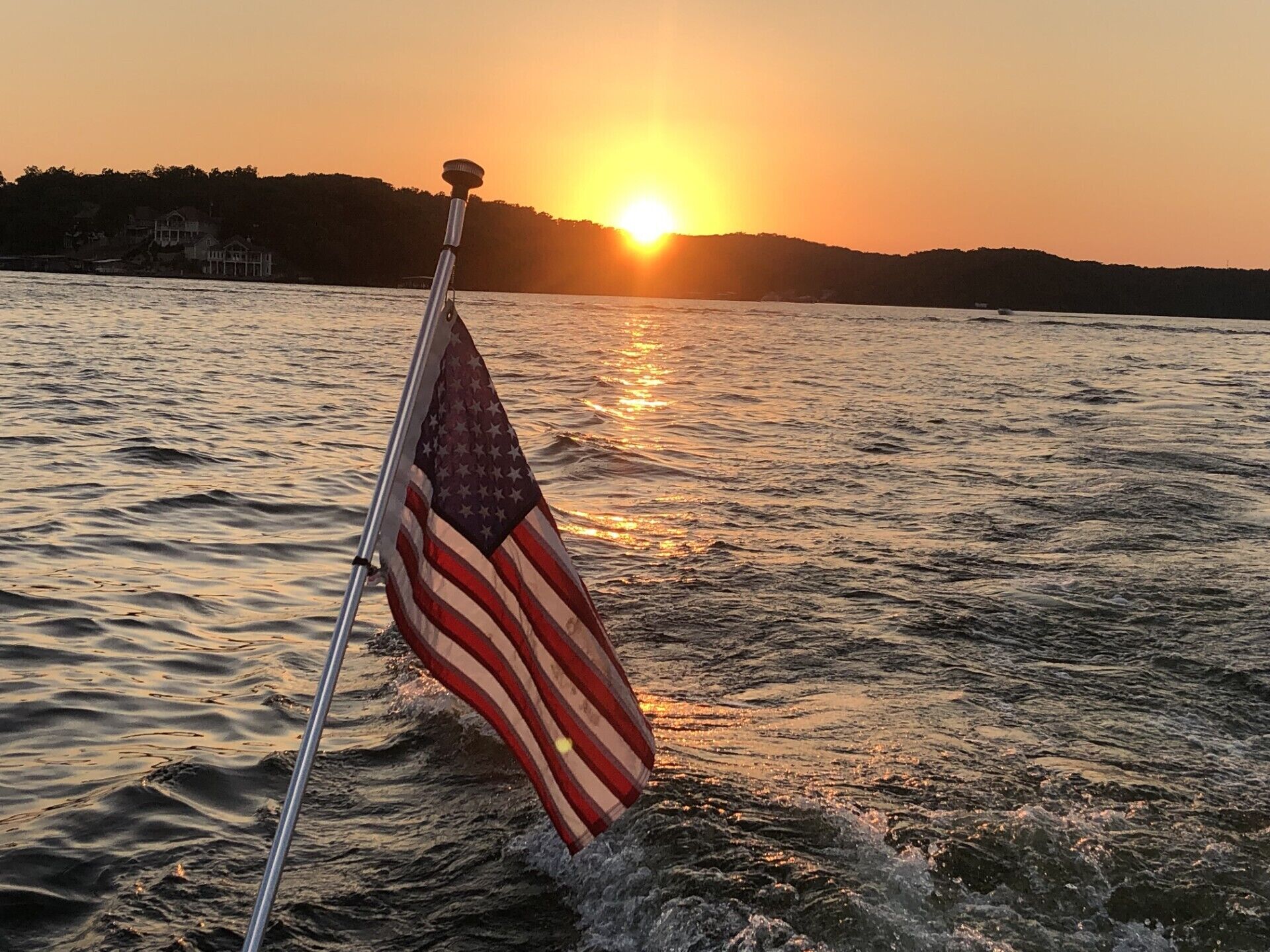 A flagpole with an American flag in front of a body of water at sunset.