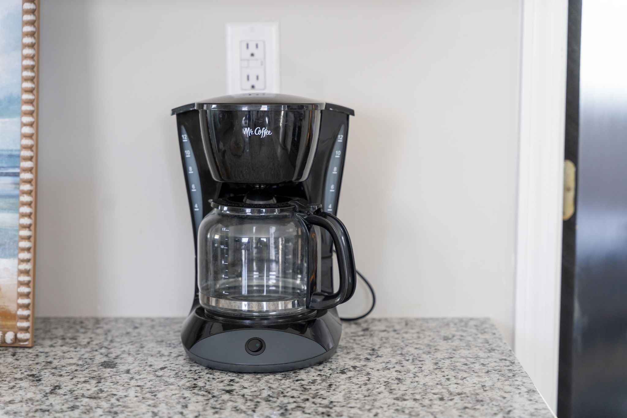 A coffee maker on a granite countertop with a plain wall and a door in the background.