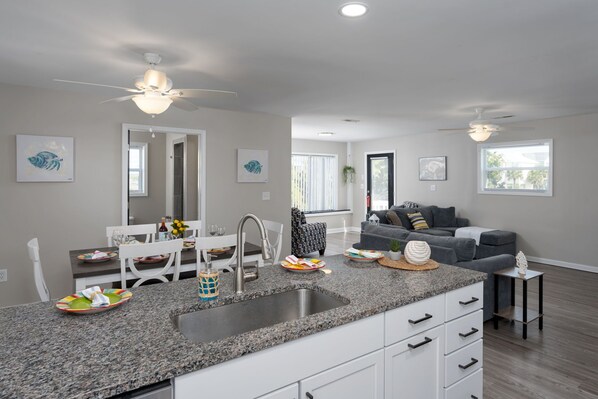 A modern kitchen with granite countertops, white cabinetry, and a built-in sink. There is a dining area with a white table and chairs, and a gray sofa in the living room.