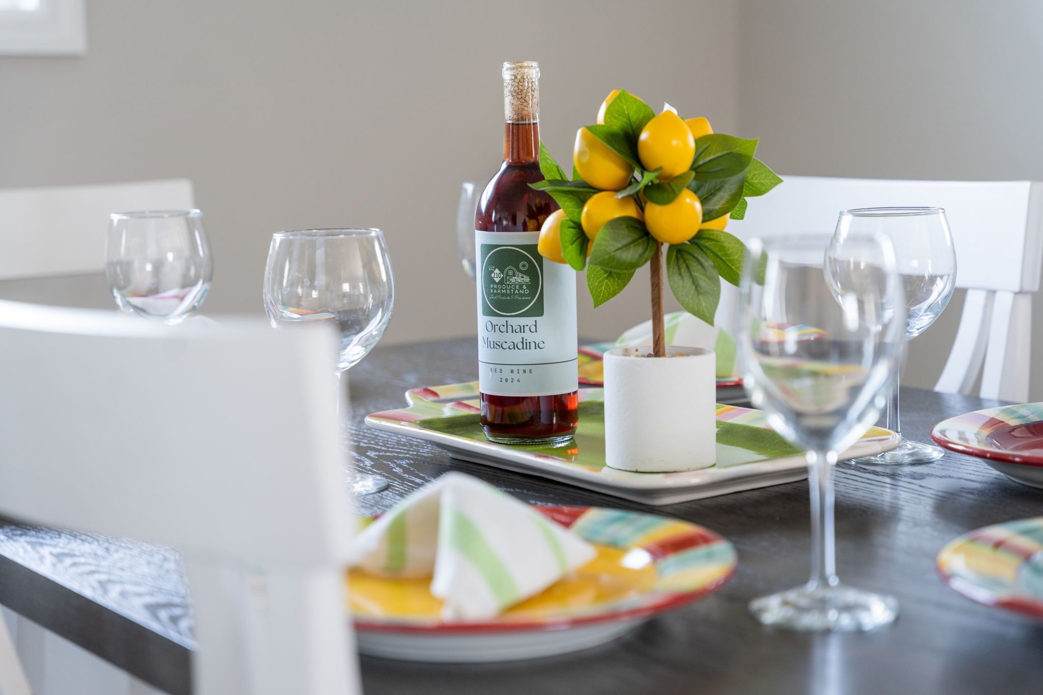 A dining table with a bottle of Orchard Muscadine red wine, a potted plant with yellow flowers, and colorful plates and glasses.