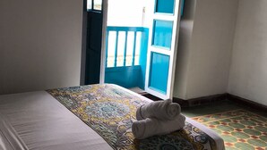 A single bed with a patterned bedspread, a rolled towel on the bed, and a view of an open balcony with blue railings. - The Best Adventure (San Gil)