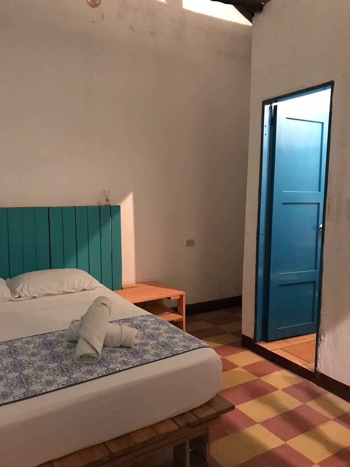A room with a bed, a small wooden table, a blue door, and a checkered floor.