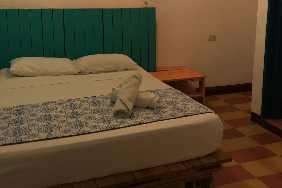 A bed with a teal headboard, white pillows, and a rolled towel on a patterned bedspread.