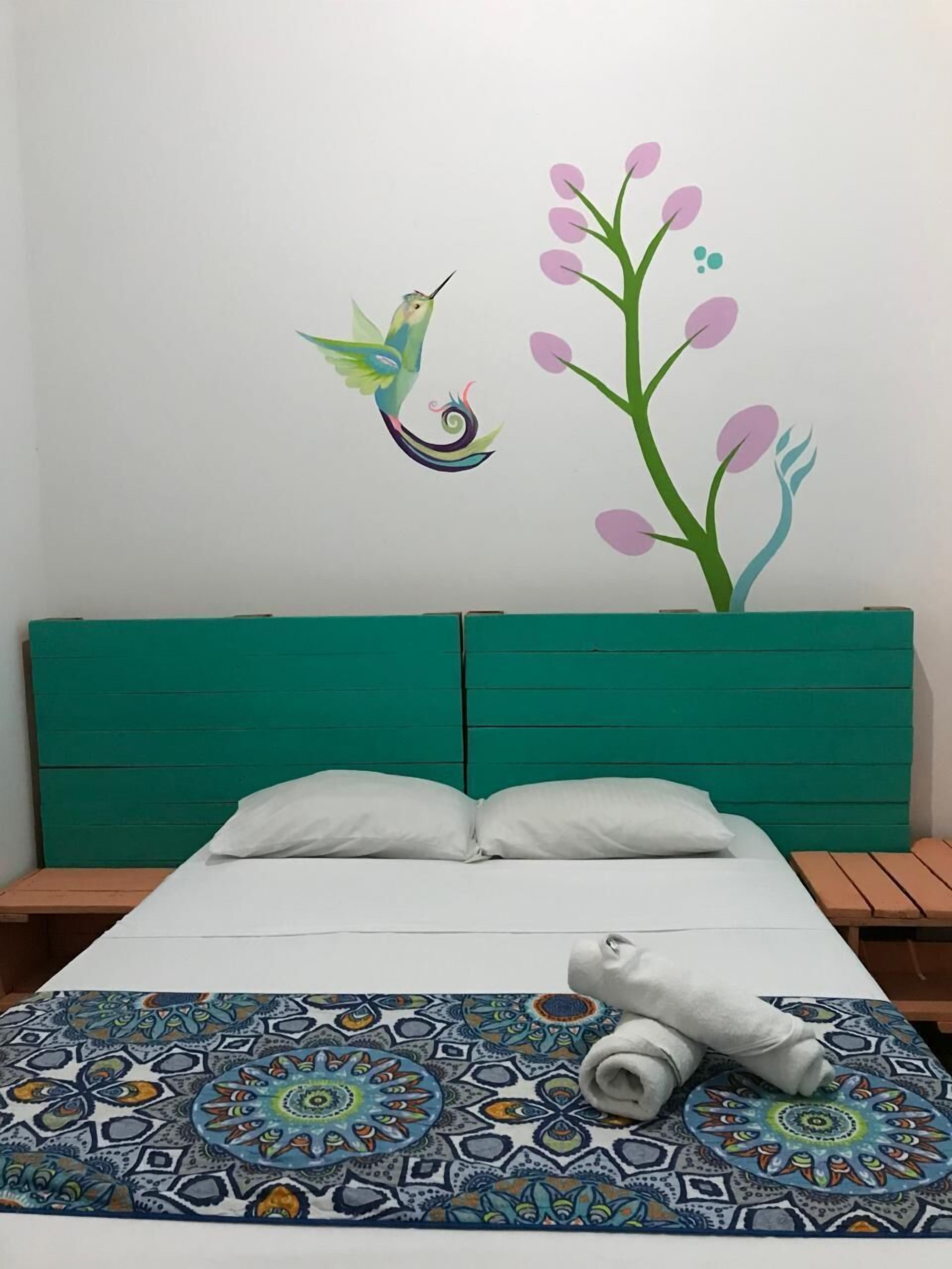 Una habitación con cama, cabecero verde, ropa de cama blanca, un tapete colorido con estampado y decoración de un pájaro y flores.