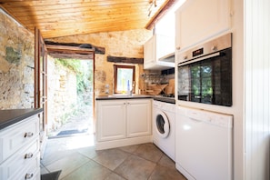 A kitchen with stone walls, wooden ceiling, white cabinets, a Bosch washing machine, and a window overlooking greenery. - The Gîte (VITRAC)
