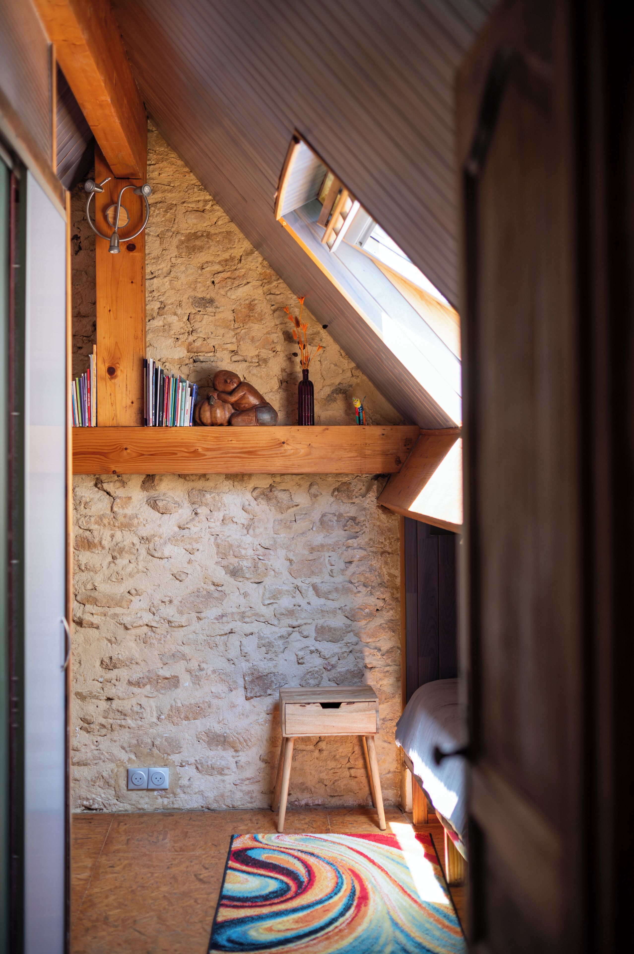 A cozy attic room with a sloped ceiling, a wooden shelf with books and a small statue, a colorful rug, and a simple wooden stool.