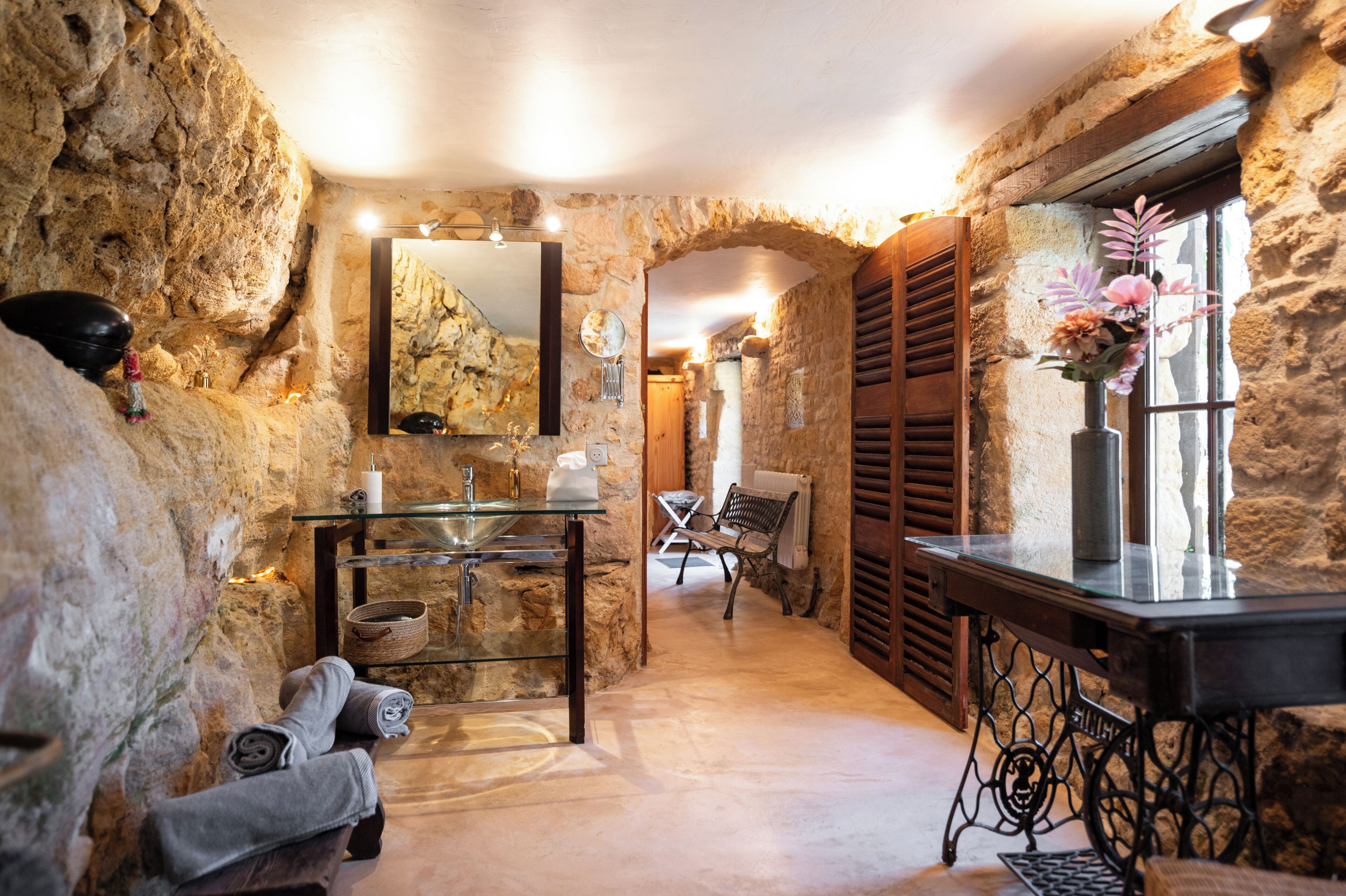 Une salle de bain aux murs en pierre, dotée d’un lavabo en verre, d’un banc et d’un miroir.
