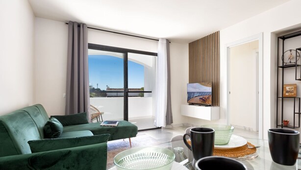 A modern living room with a green sofa, a dining table with glassware, and a large window offering a view of the sea. - Delfin | Pool - terrace - golf (Mijas Costa)