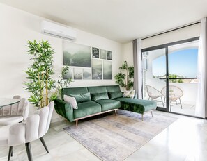 A modern living room with a green sofa, a dining table, and chairs. There are several plants, a large window with a view, and a sliding glass door leading to an outdoor area.