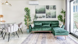 A modern living room with a green sectional sofa, a glass dining table with chairs, a large window with a view, and a wall-mounted air conditioner. - Delfin | Pool - terrace - golf (Mijas Costa)