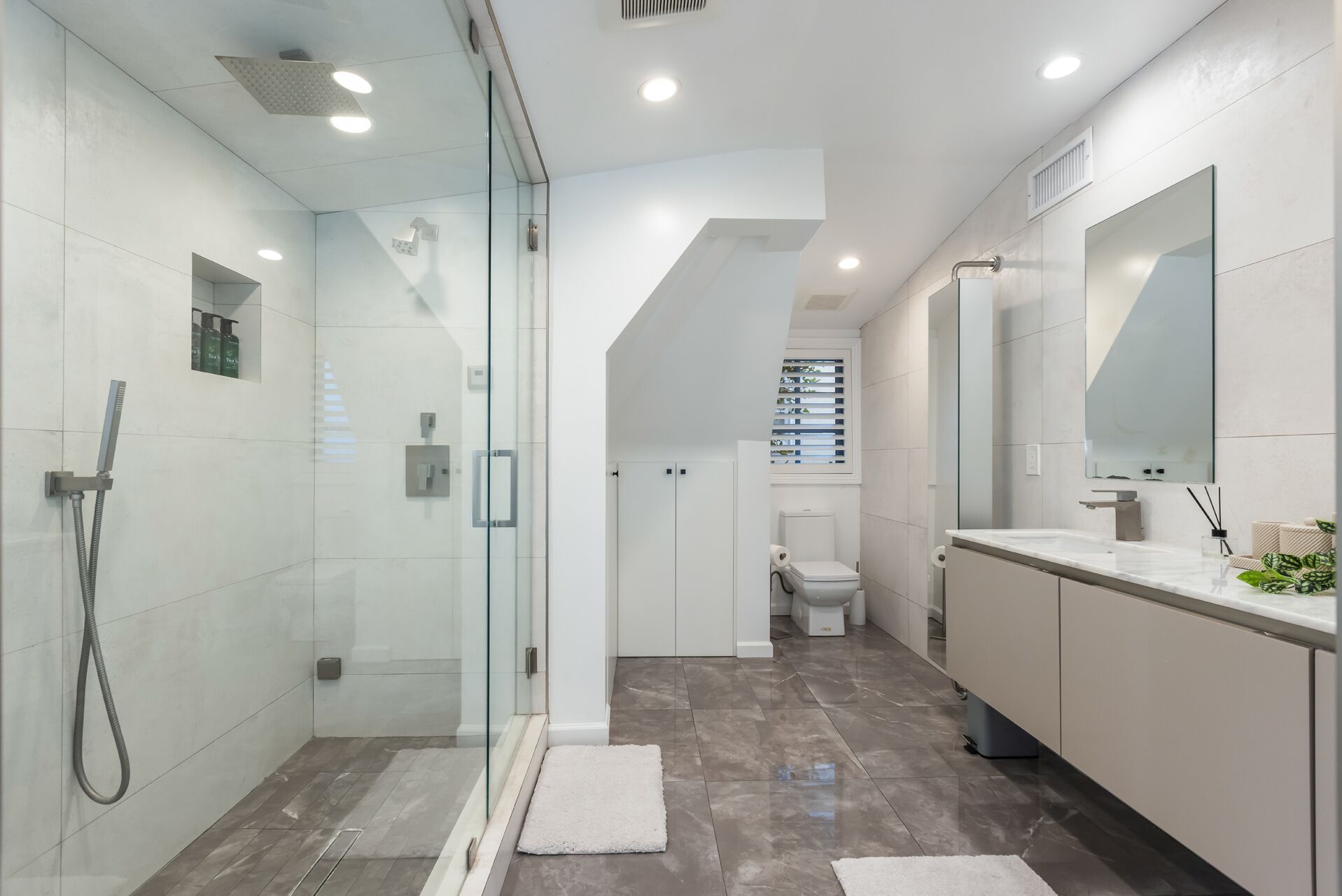 Un baño moderno con ducha envuelta en vidrio, un tocador blanco con un espejo grande y un inodoro visible al fondo.