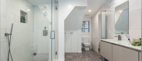 Un baño moderno con ducha envuelta en vidrio, un tocador blanco con un espejo grande y un inodoro visible al fondo.