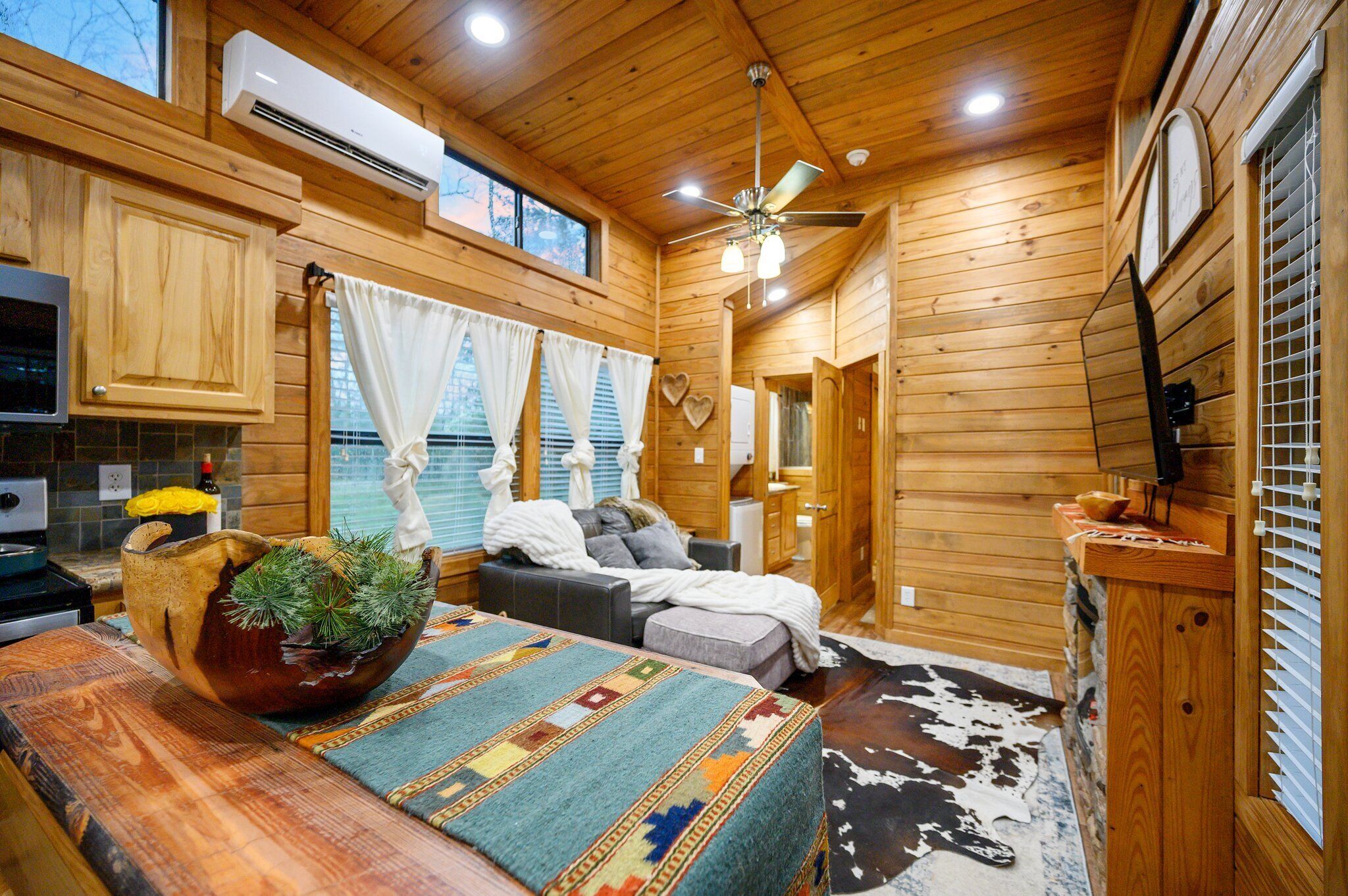 L’intérieur chaleureux d’un chalet avec des murs en bois, un ventilateur de plafond, un grand saladier en bois rempli de verdure, un lit recouvert d’une couverture à motifs et un tapis en peau de vache.
