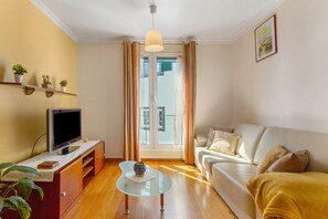 Een woonkamer met een bank, een glazen salontafel, een tv op een houten dressoir, en een plant.