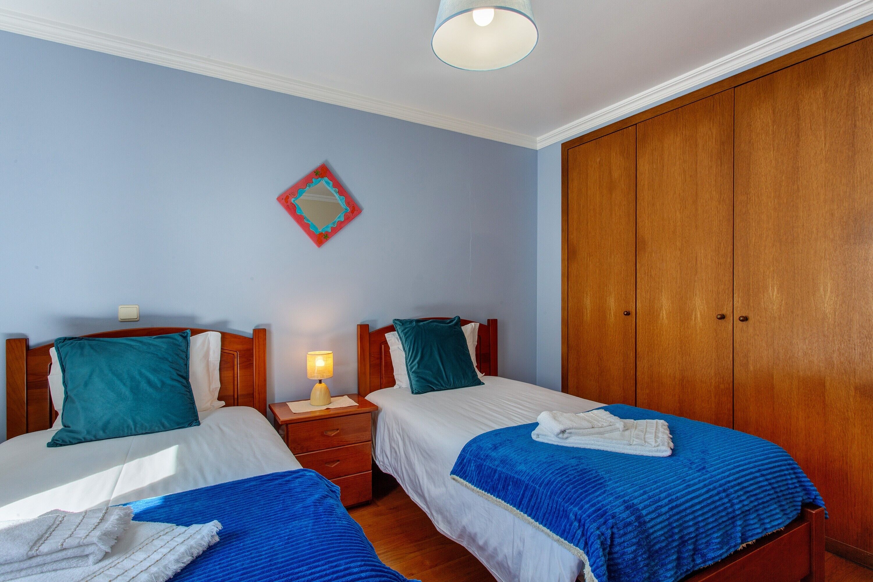 A hotel room with two beds, a wooden wardrobe, a nightstand with a lamp, and a wall decoration.