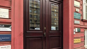 Un edificio rojo con una puerta doble marrón oscuro, varios letreros incluyendo 'SALON SPA' y 'KANCELARIA ADWOKACKA', y el reflejo de un edificio en los paneles de vidrio.