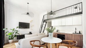 A modern living room with a sofa, two wooden chairs, a white table, a potted plant, and a TV mounted on the wall.