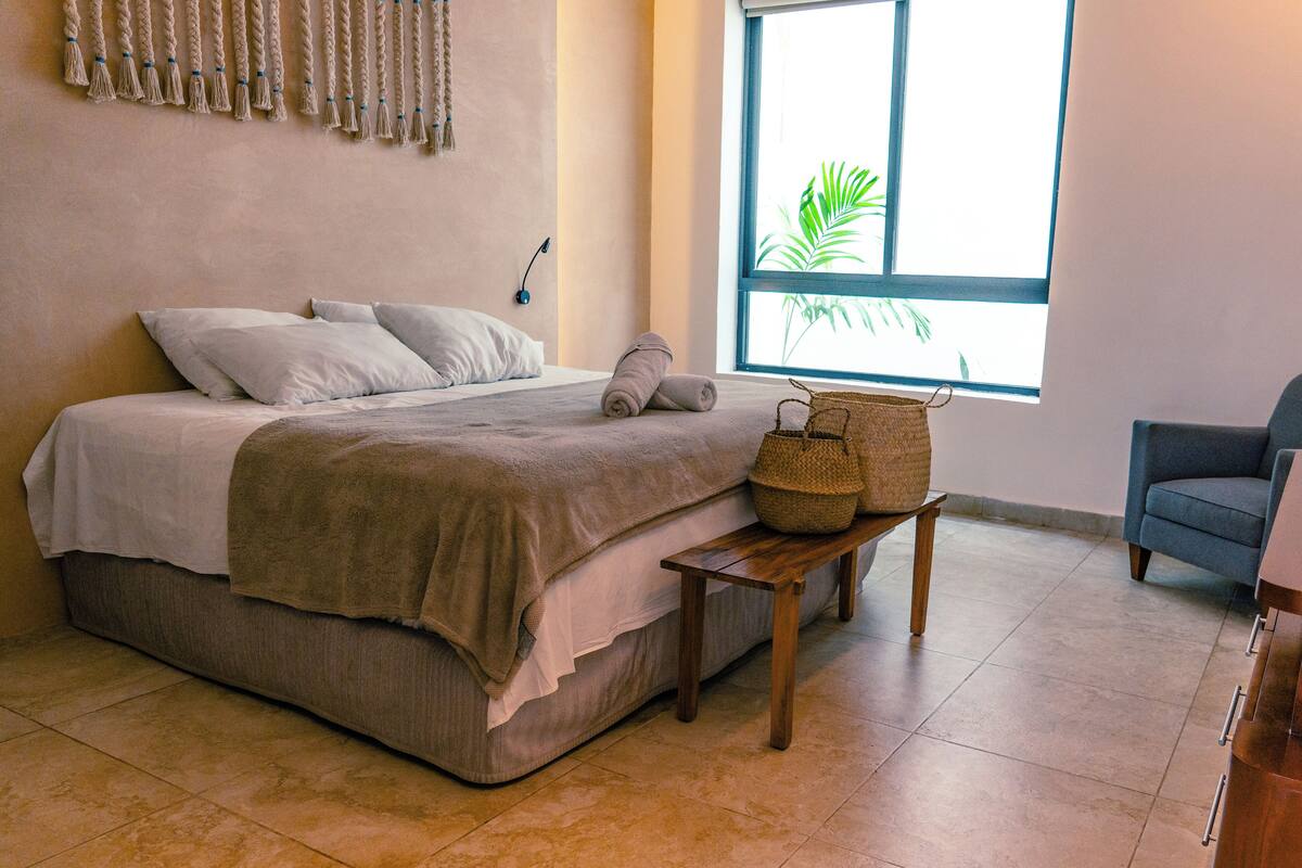 A bedroom with a bed, a bedside table with baskets, a chair, and a window with a plant view.