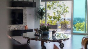Un salon moderne avec une table basse en verre, un téléviseur à écran plat fixé à un mur en pierre et une grande fenêtre donnant sur un paysage verdoyant.