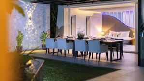 Un espace extérieur moderne aménagé pour les repas, avec un mur en pierre et des plantes en pot, un coin salon bien éclairé avec un canapé et une table, et une vue sur l’intérieur.