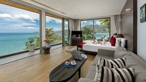 Une chambre d’hôtel moderne dotée d’un grand lit, d’un canapé, d’une télévision et d’une table à manger. La chambre dispose de grandes fenêtres offrant une vue sur la mer et les collines.