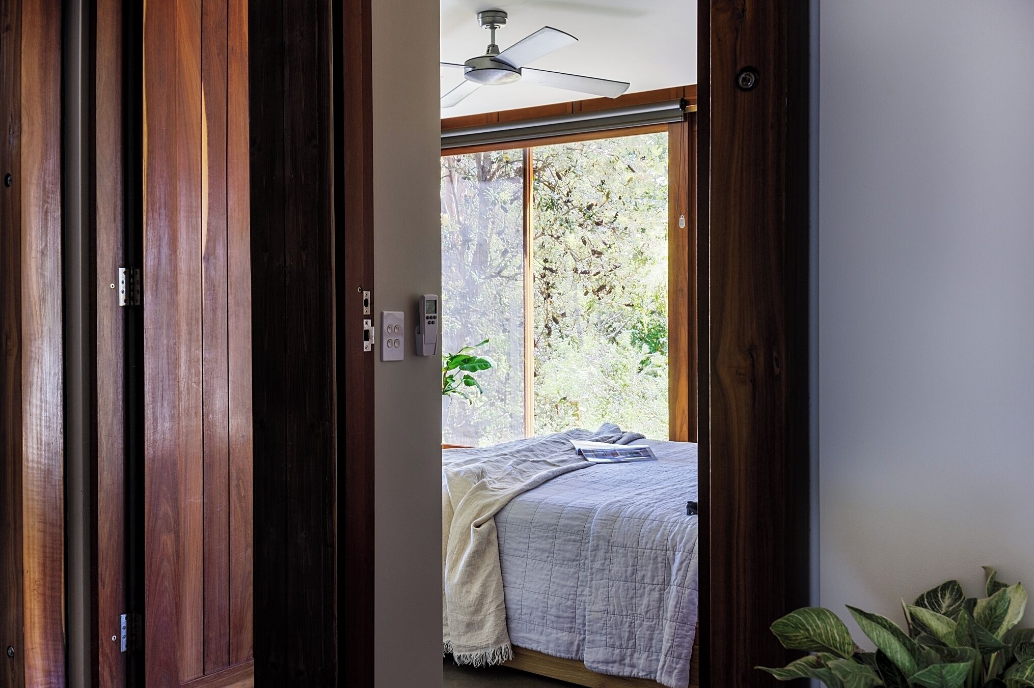 Une chambre à coucher avec un lit, un ventilateur de plafond et une vue sur les arbres, visible à travers une porte coulissante.