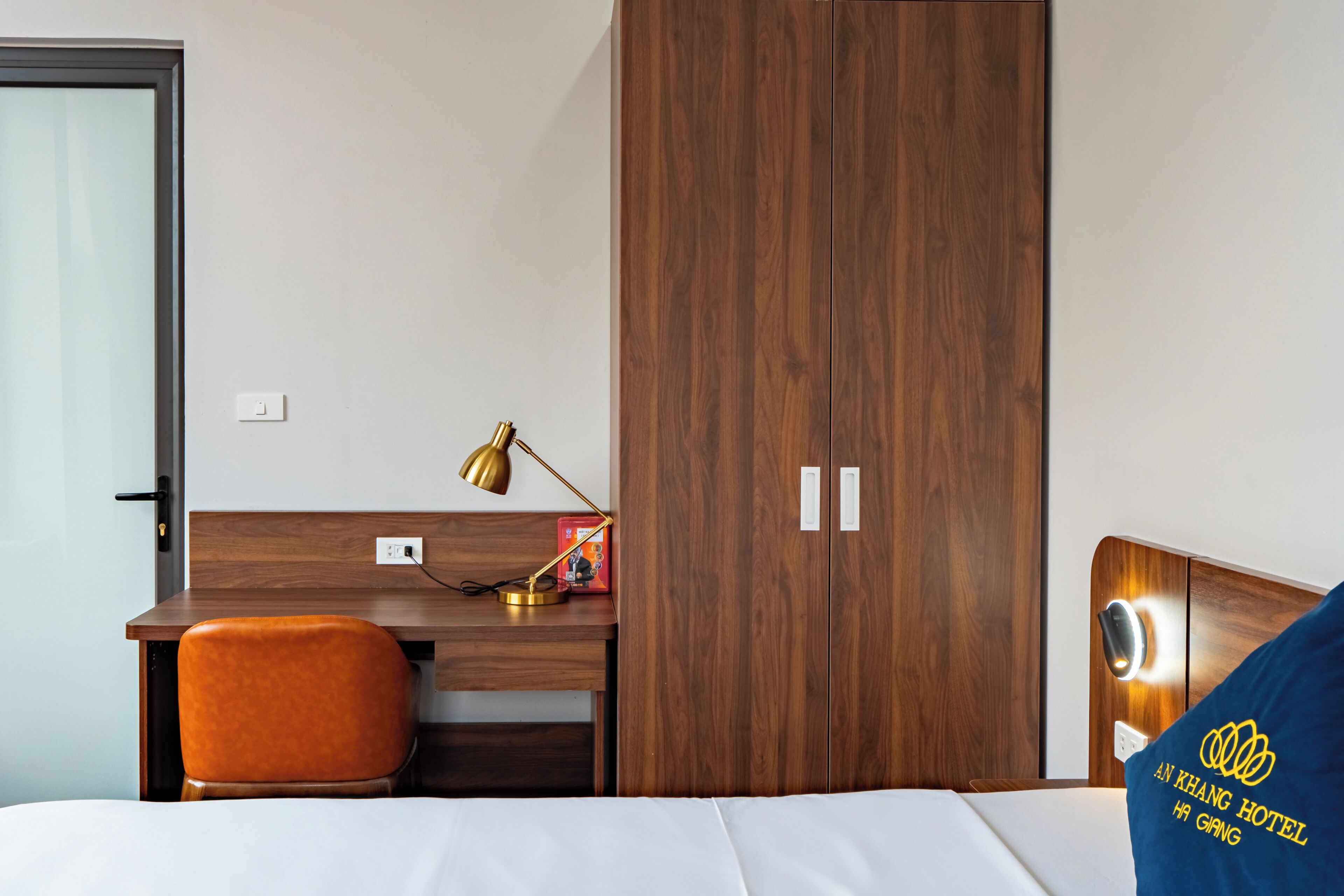 A hotel room with a wooden desk, an orange chair, a bed with a blue pillow, and a wardrobe.