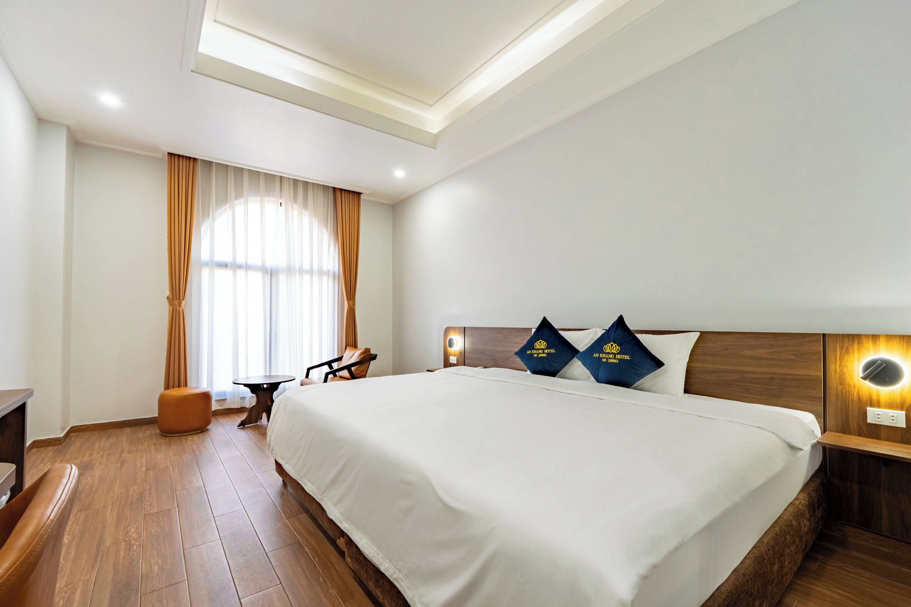 A hotel room with a large bed, a desk, a chair, a small ottoman, and a window with curtains.