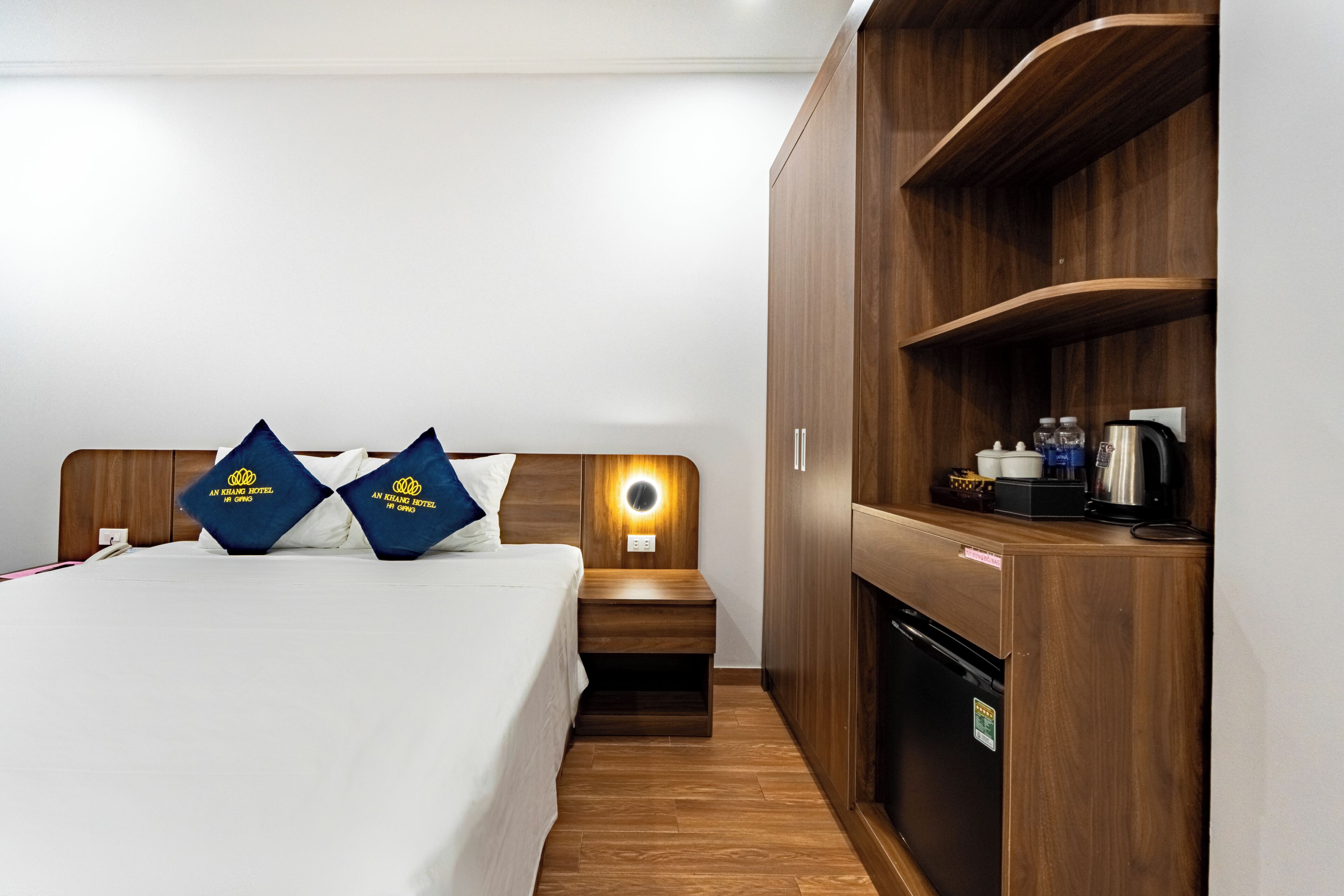 Habitación de hotel con cama, mesita de noche y una estantería de madera con cafetera.