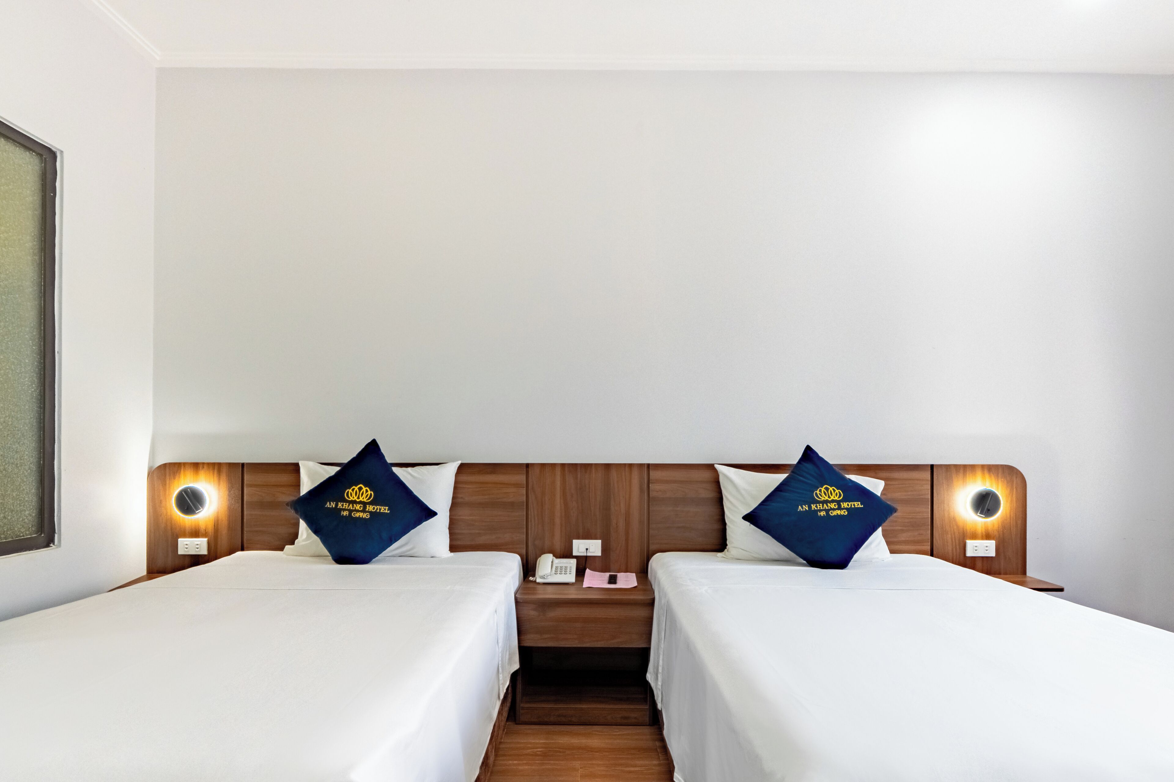 A hotel room with two beds, a nightstand with a phone and a lamp, and a wall-mounted light fixture.