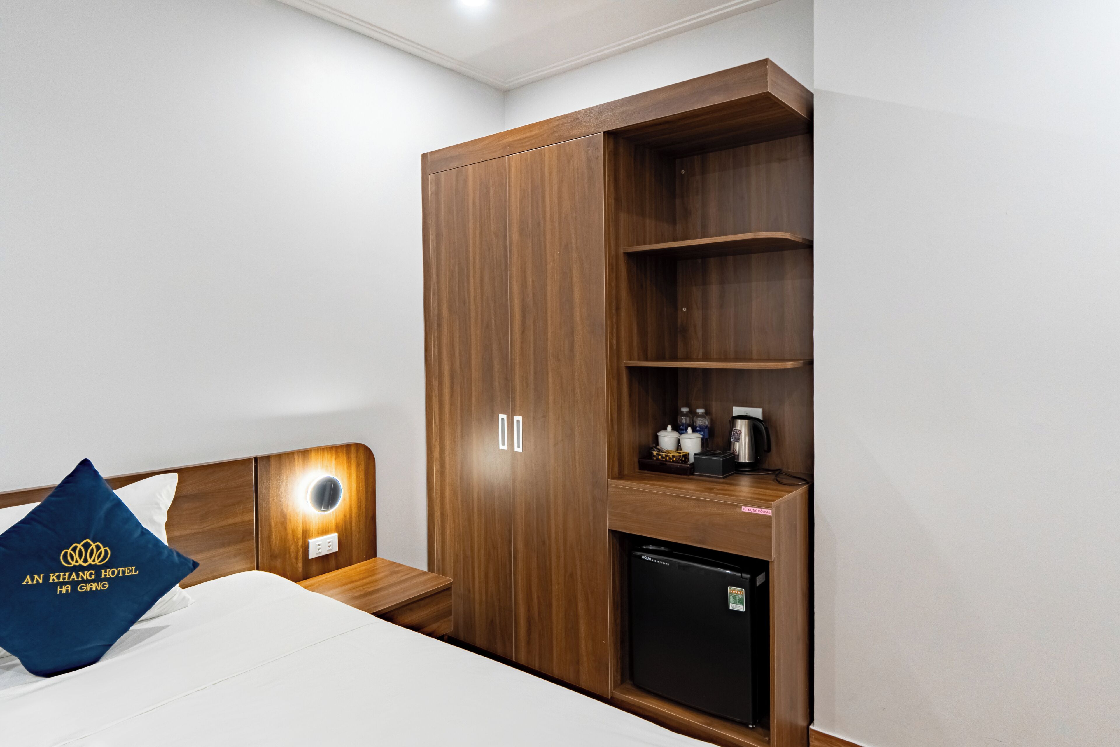 Una habitación de hotel con cama, armario de madera, refrigerador pequeño y mesita de noche con lámpara.