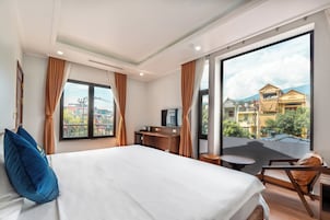 Habitación de hotel con una cama grande, un escritorio, una silla y vistas a edificios a través de un ventanal.