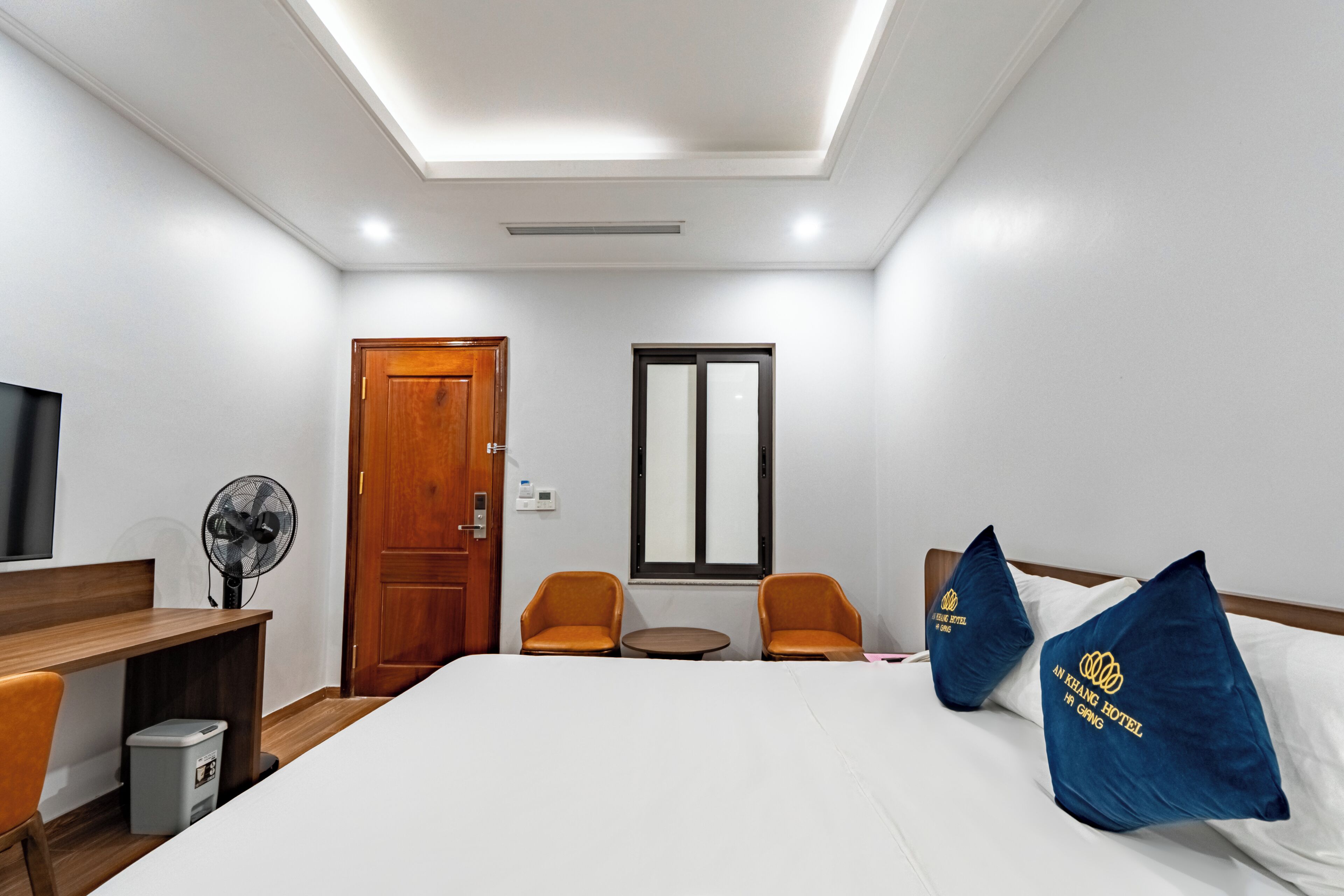 Una habitación de hotel con una cama, dos sillas, una mesita, un televisor, un ventilador y una puerta.