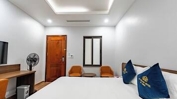 Una habitación de hotel con una cama, dos sillas, una mesita, un televisor, un ventilador y una puerta.