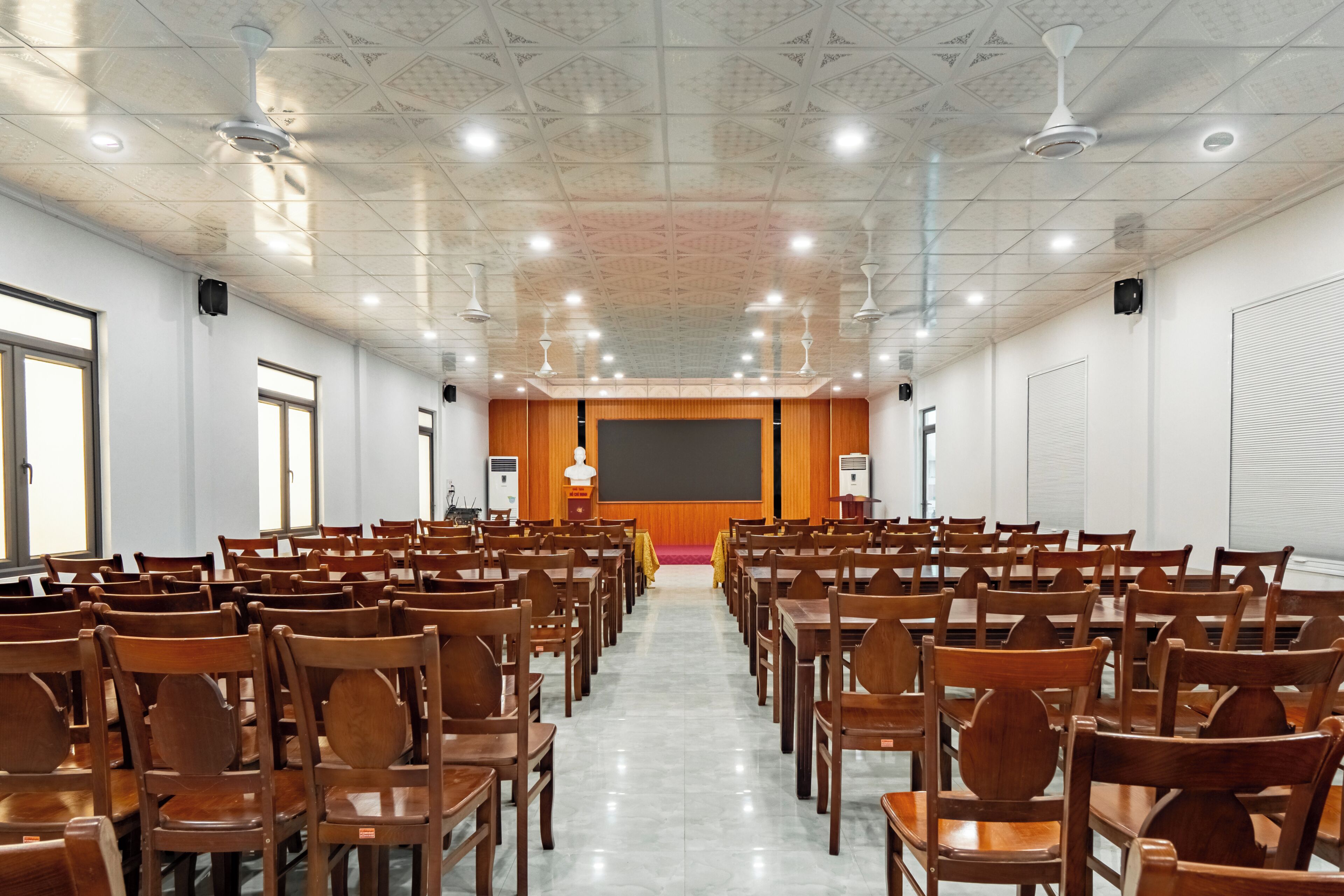 Un salón de conferencias con filas de sillas de madera, una pantalla grande y ventiladores de techo.
