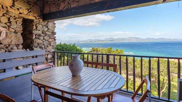 A wooden table and chairs on a balcony with a stone wall, overlooking a coastal view. - Apartment 'Casa Vacanze Ribes' with Sea View, Shared Pool and Private Terrace (olbia)