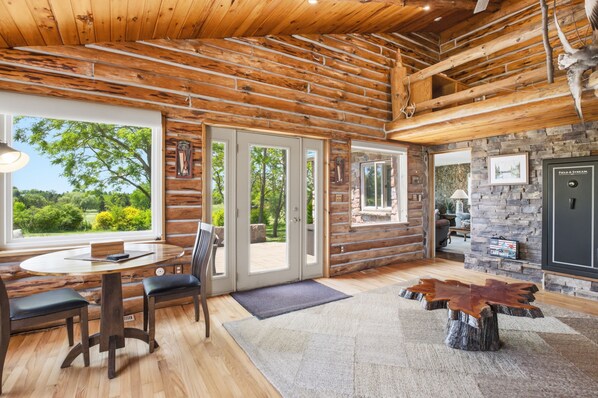 A log cabin interior with a dining area, stone fireplace, and large windows.