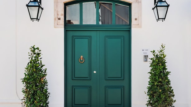 Eine grüne Tür mit kleinem Fenster darüber, gesäumt von zwei Blumentöpfen.