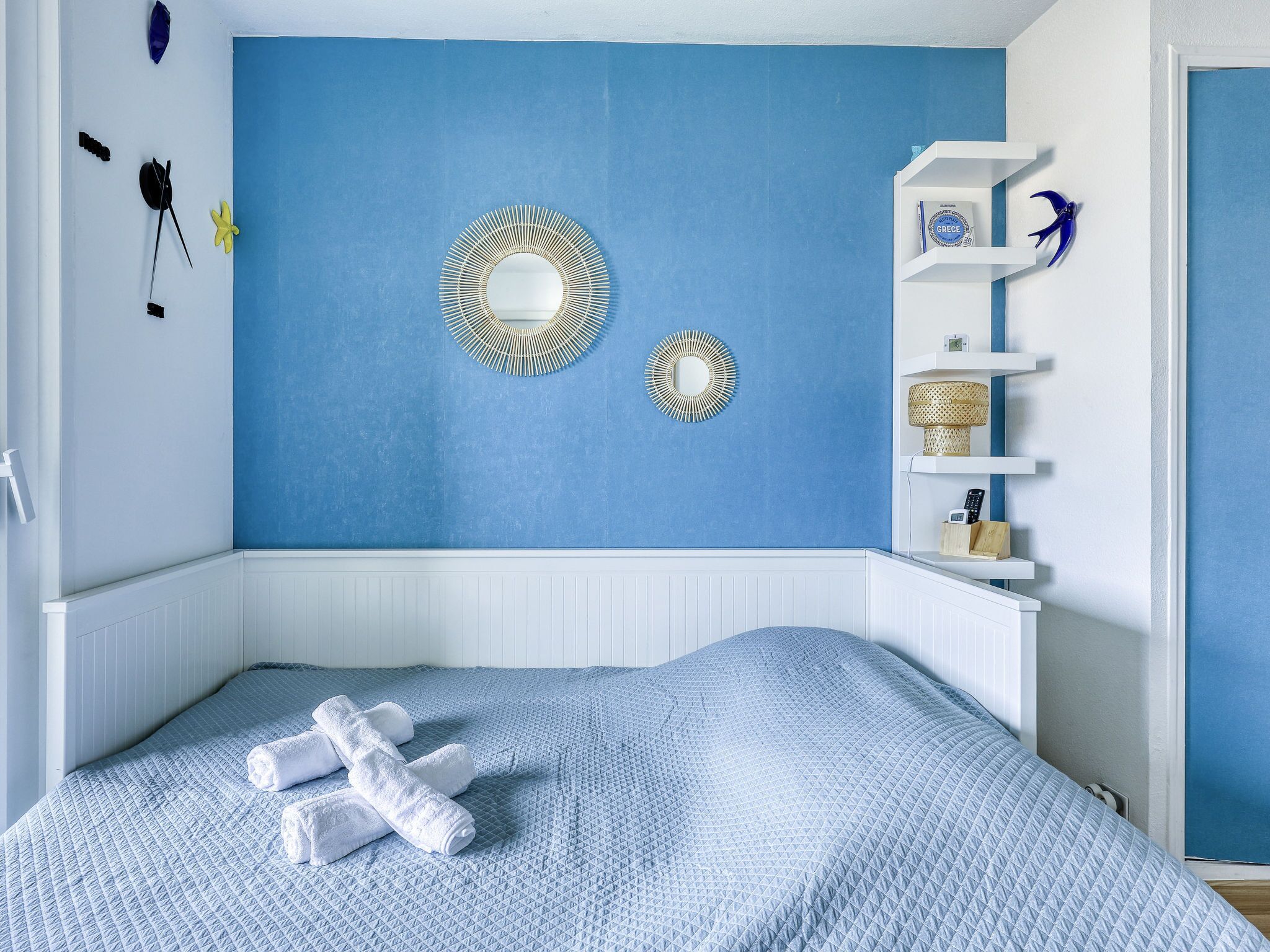 Un dormitorio con una pared azul, una cama blanca y una estantería blanca con objetos decorativos.