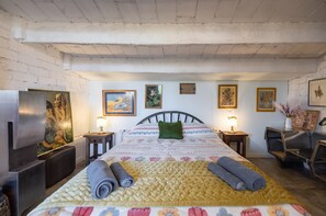 A bedroom with a bed, pillows, a nightstand, a lamp, a chair, and artwork on the walls.