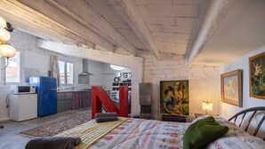 A bedroom with a bed, a large red letter 'N', a painting, and a kitchenette in the background.