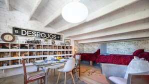 A room with a white brick wall, a sign reading '15 DESPOJOS,' a dining table with chairs, a sofa, and a patterned rug.